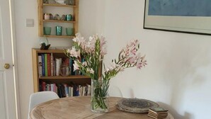 Ein runder Holztisch mit zwei weißen Stühlen, eine Vase mit Blumen und ein Bücherregal im Hintergrund.