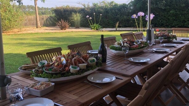 Un espace de restauration en plein air avec une table en bois dressée pour un repas, entourée de chaises, et un grand parasol offrant de l’ombre.