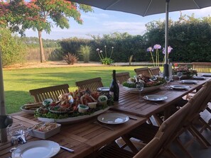 An outdoor dining area with a wooden table set for a meal, surrounded by chairs, and a large umbrella providing shade. - Spacious house on a wooded lot: swimming pool, hot tub, and tennis court (Saint-Geours-de-Maremne)