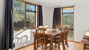 A dining area with a wooden table and chairs, large windows with curtains, and a view of greenery outside.