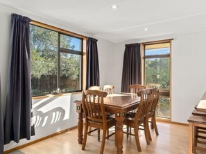 A dining area with a wooden table and chairs, large windows with curtains, and a view of greenery outside. - Holiday Heaven - Cromwell (Cromwell)