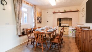 A dining area with a wooden table set for six, surrounded by wooden chairs. A kitchen with a stove and cabinets is visible in the background. - The Vale (Beaworthy)