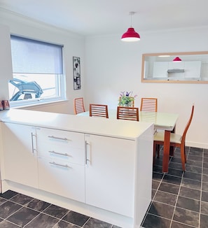 A compact kitchen with a central island, white cabinetry, a window with blinds, a pendant light, and a dining area with chairs and a table. - Peaceful bungalow, super close to birnam oak and hiking trails. (Dunkeld)