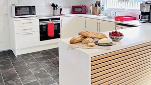 Une cuisine moderne avec des meubles blancs, un luminaire suspendu rouge, un four à micro-ondes, un grille-pain, une machine à café et un plan de travail en bois sur lequel sont posés du pain et des fruits.