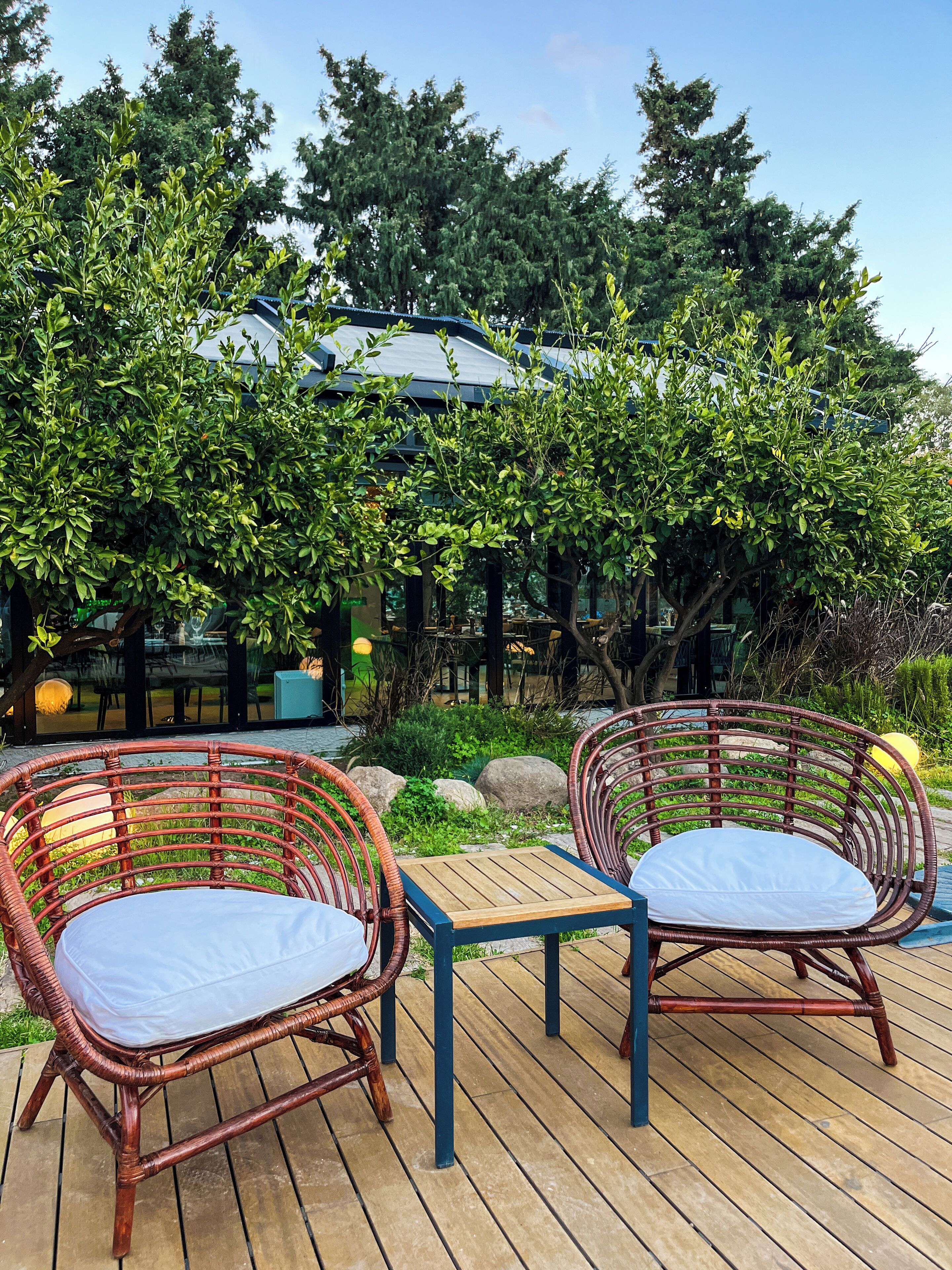 Des chaises en osier avec des coussins blancs, placées sur une terrasse en bois, entourées de verdure et d’un bâtiment doté d’un toit bleu.