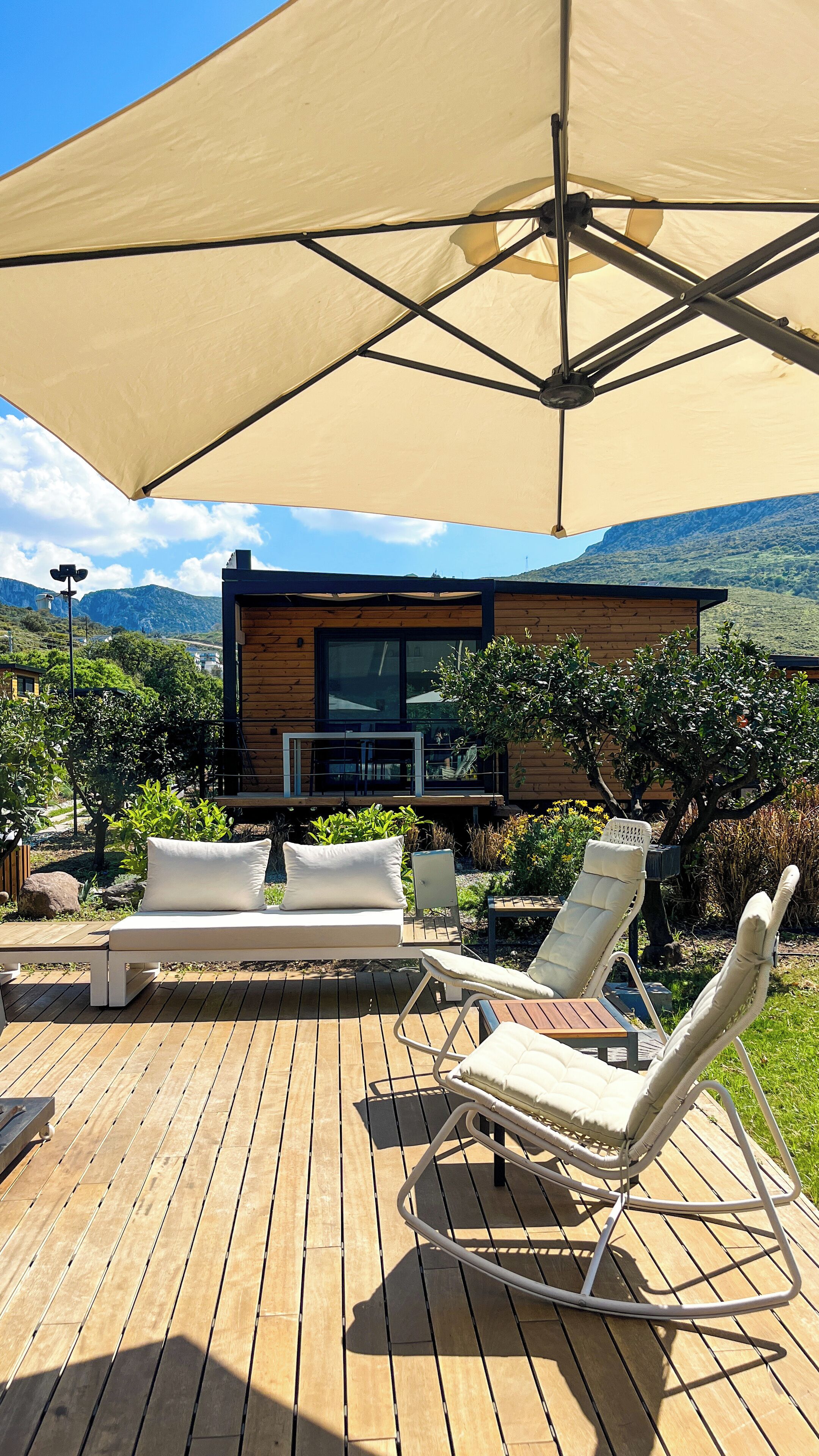 Una terraza al aire libre con piso de madera, un amplio toldo color beige y dos sillones blancos.