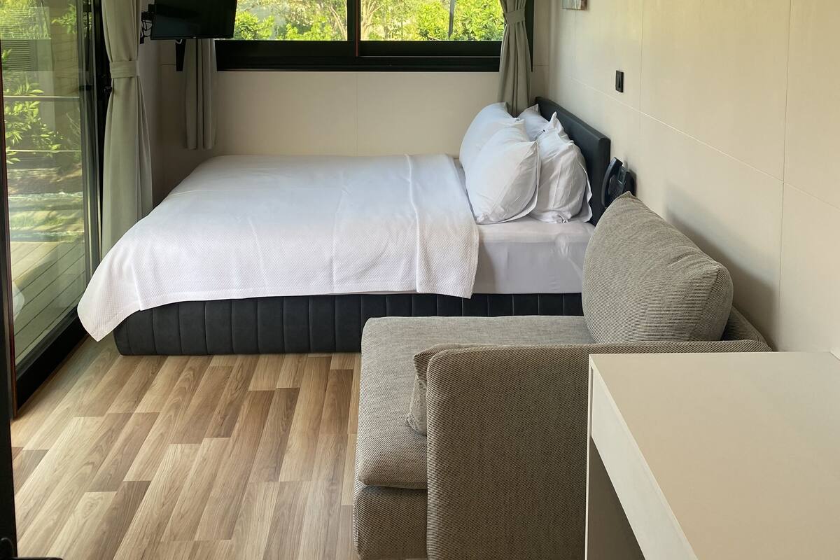 Un dormitorio con cama, una silla, una mesita, un ventanal con cortinas, un equipo de aire acondicionado y vistas a zonas verdes.