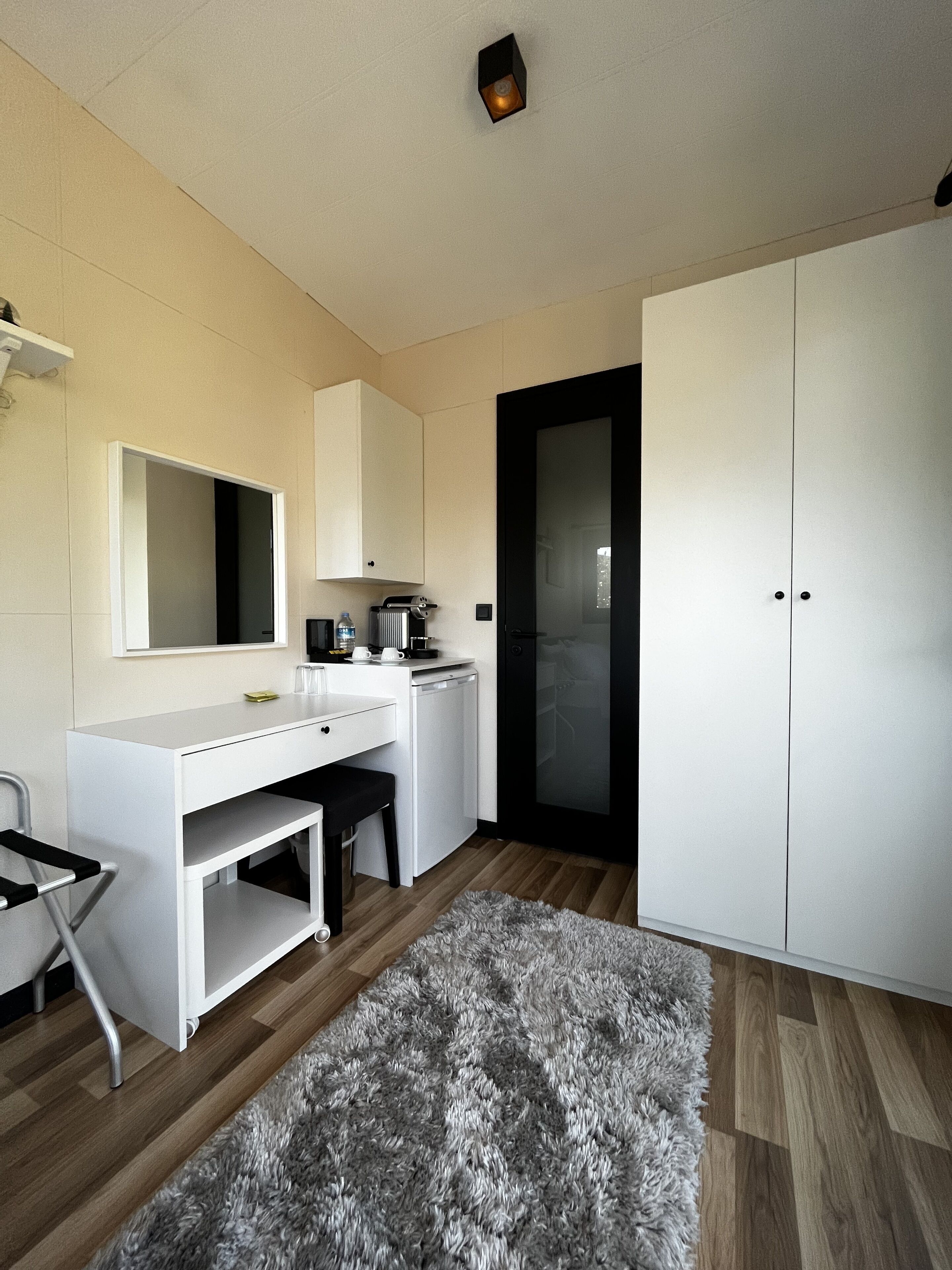 Et moderne, minimalistisk soveværelse med et hvidt skab, et skrivebord med stol, et lille køleskab og en blød måtte på trægulvet.