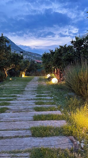 A stone pathway leading through a garden with grass and plants, surrounded by trees and a mountain in the background. - Tiny Town Kuyucak (Karaburun)