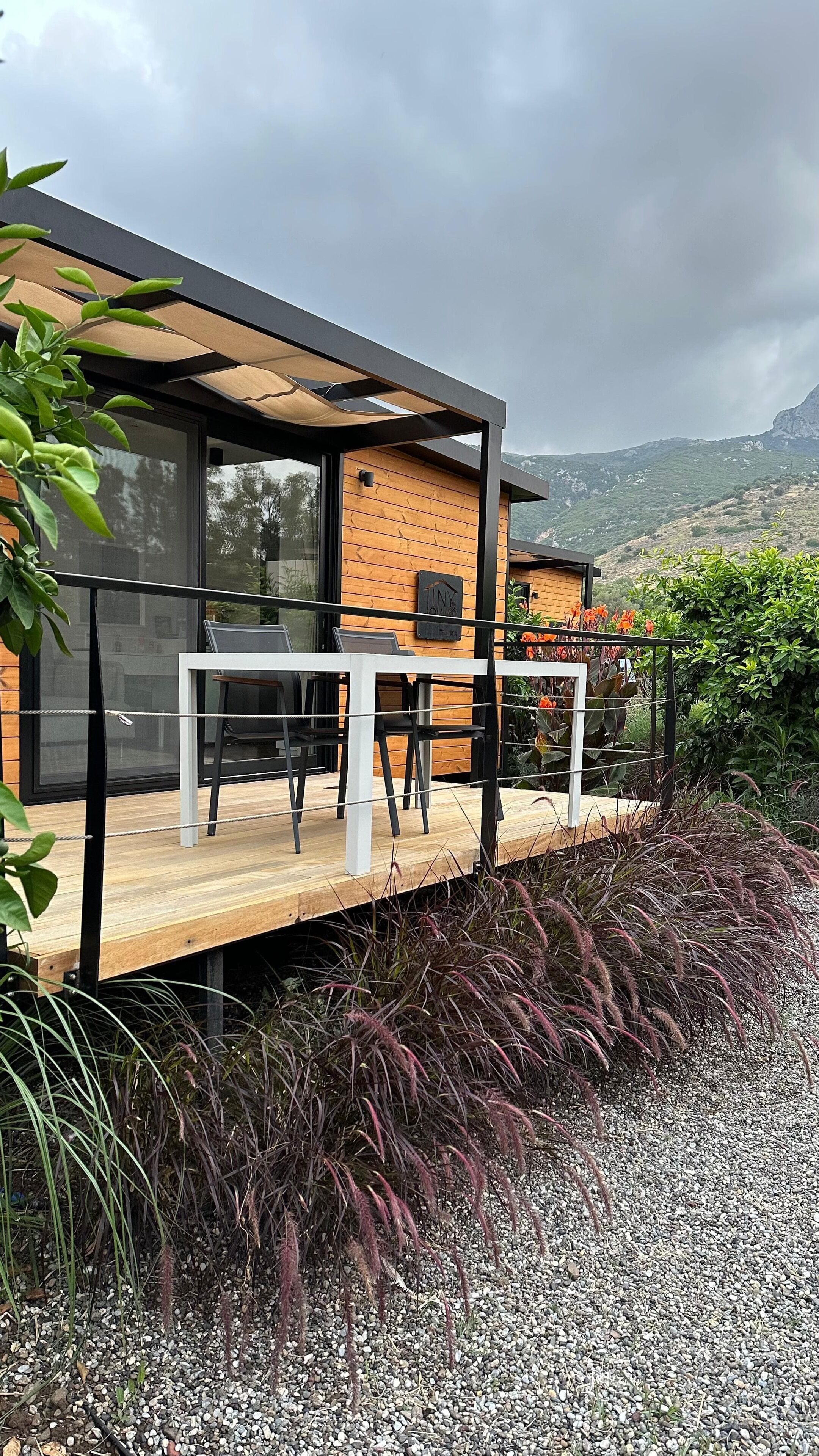 A modern outdoor seating area with a wooden deck, black chairs, and a glass railing overlooking a mountainous landscape.
