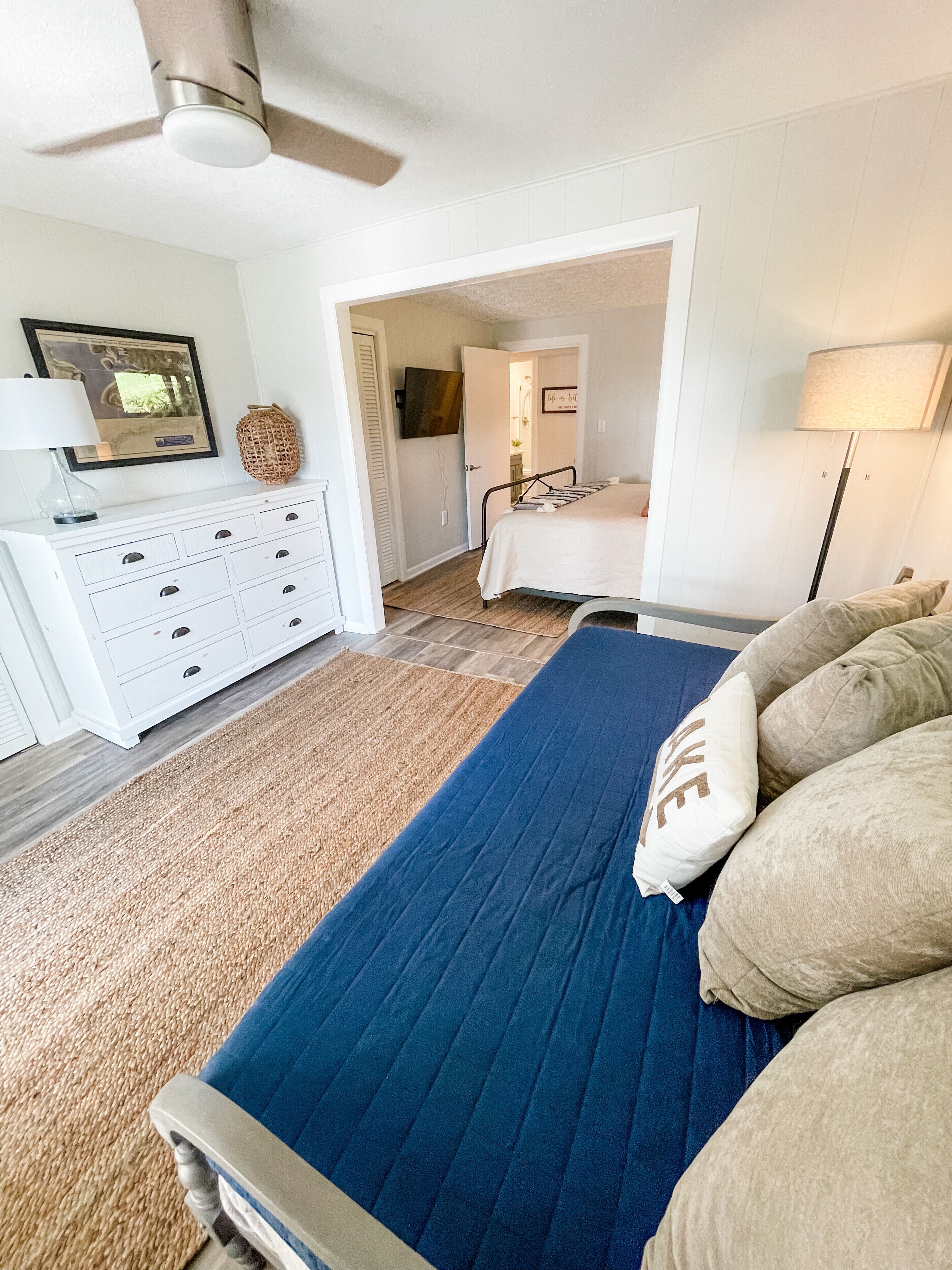 A bedroom with a bed, a dresser, a ceiling fan, and a door leading to another room.