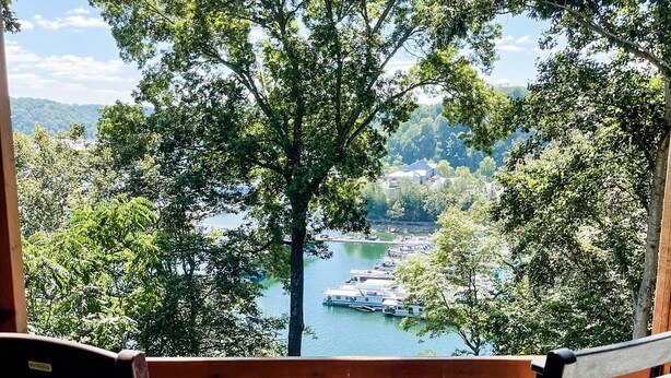 A wooden deck with two chairs facing a lake with boats, surrounded by trees and a view of a hilly landscape.