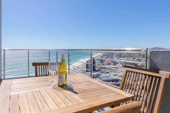 A wooden table with a bottle of wine and a glass on a balcony overlooking a beach and cityscape.