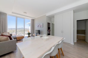 A modern living room with a large window, a dining area with a table and chairs, and a kitchen area.