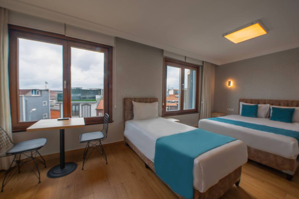 A hotel room with two beds, a table, and chairs. There are large windows offering a view of buildings outside.