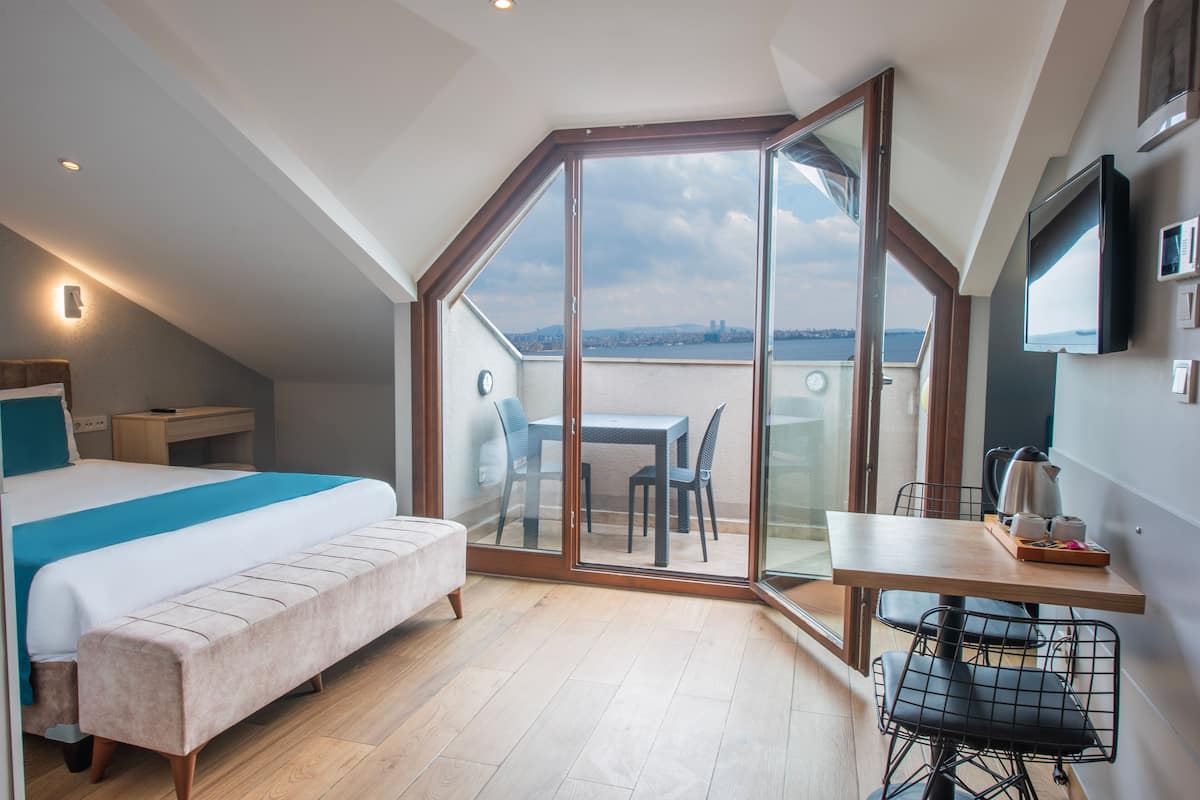 A modern hotel room with a large bed, a bench, a dining table with chairs, a TV, and a balcony with a view of the city.