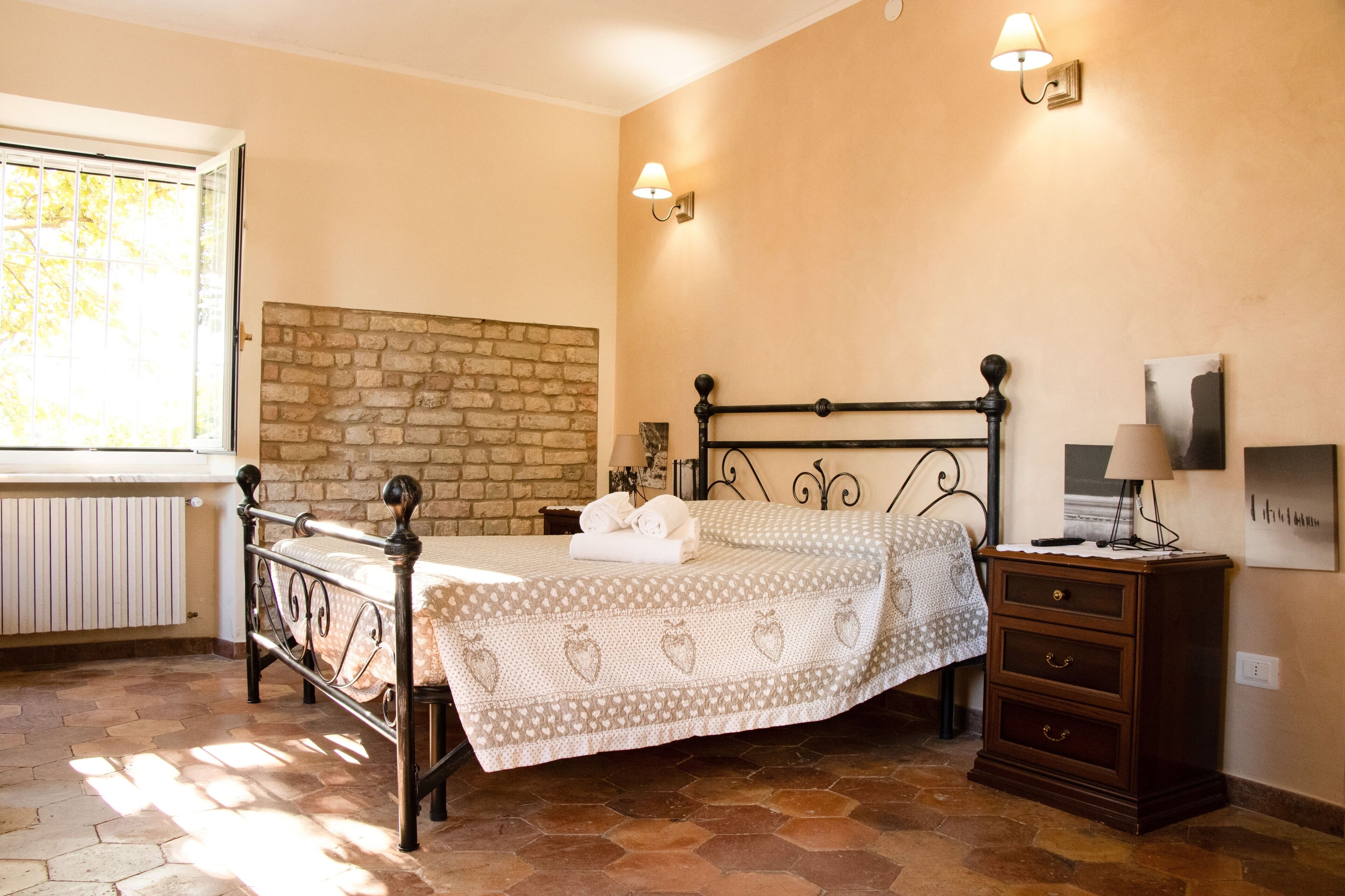 A bedroom with a brick wall, a bed with a patterned bedspread, a nightstand with a lamp, and a window with blinds.