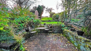 Un patio giardino con pavimento in pietra, tavoli e sedie in metallo, circondato da piante rigogliose e un piccolo stagno.
