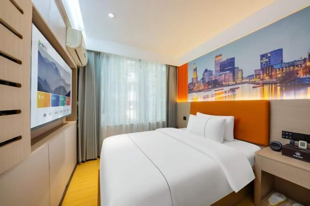 A hotel room with a bed, a cityscape painting, a wall-mounted TV, and a wooden wardrobe.