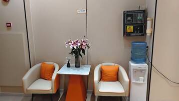 A room with two chairs, a small table with a vase of flowers, a water dispenser, and a control panel on the wall.