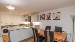 A modern kitchen with a central island, dining table, and chairs. There are framed pictures on the wall, a floor lamp, and a washing machine.