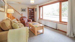 A cozy living room with a sofa, a wooden coffee table, a bookshelf, and a large window with curtains. - Iola (Inverness)