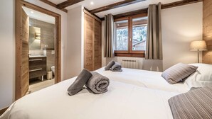 Une chambre d’hôtel avec deux lits, des poutres en bois au plafond, une fenêtre avec des rideaux et une salle de bain visible à travers une porte ouverte.