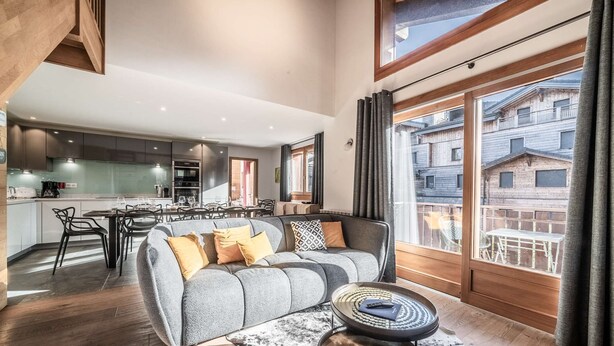 Un salon moderne avec un canapé gris, une table basse et une vue sur un balcon bordé de bâtiments en bois.