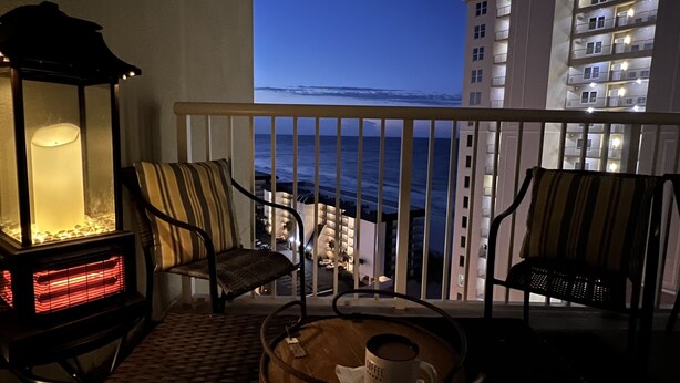 A balcony with two chairs, a table with a coffee cup, and a lit table lamp. The view includes a body of water and a building with illuminated windows.