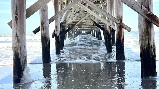 Eine hölzerne Steg, der sich in einen sandigen Strand erstreckt, wobei Wellen an seinem Fuß schlagen.