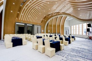 Uma grande sala de conferências com fileiras de mesas e cadeiras, teto alto com design exclusivo e janelas amplas.
