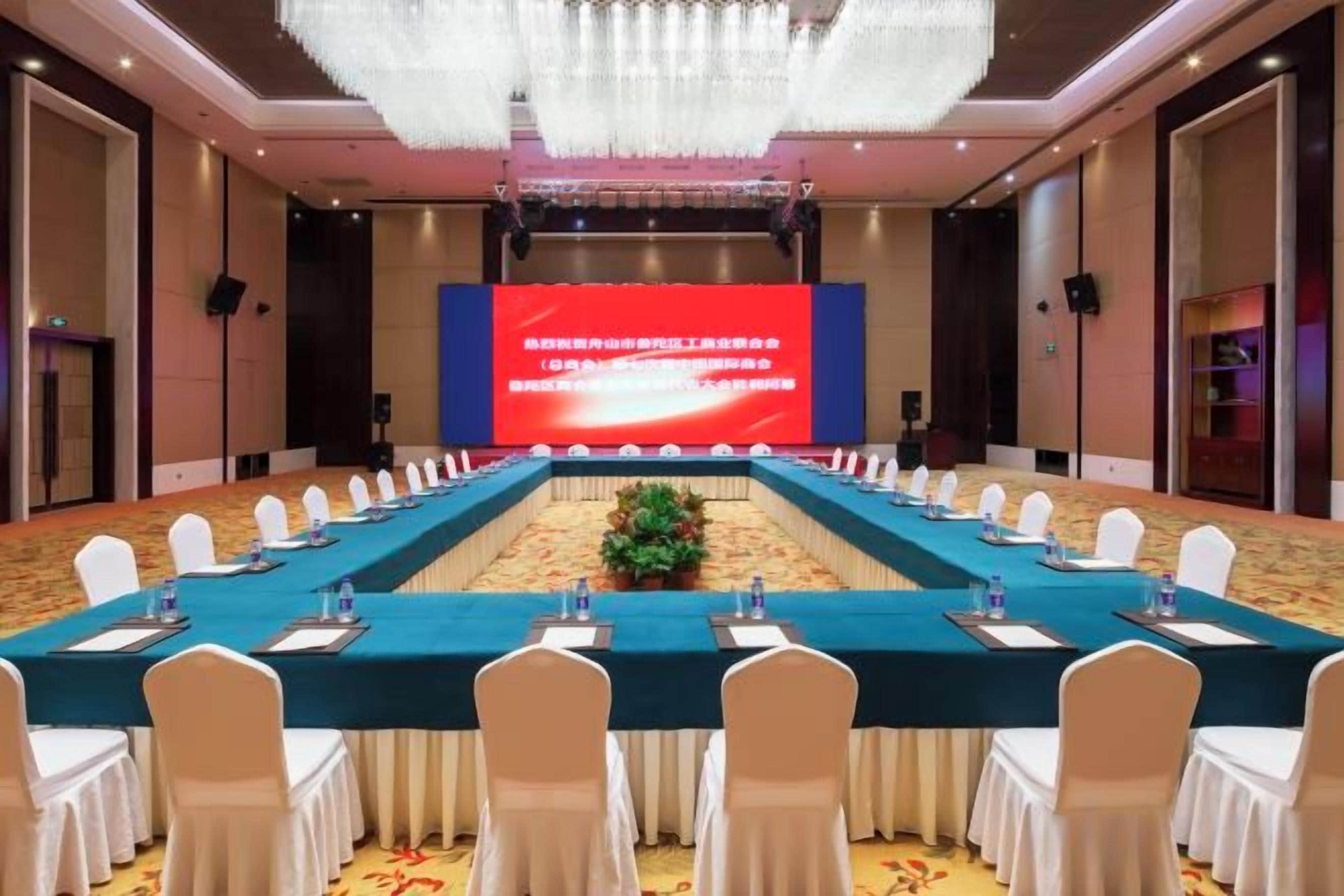 A conference room with a long table, white chairs, and a large screen at the front.