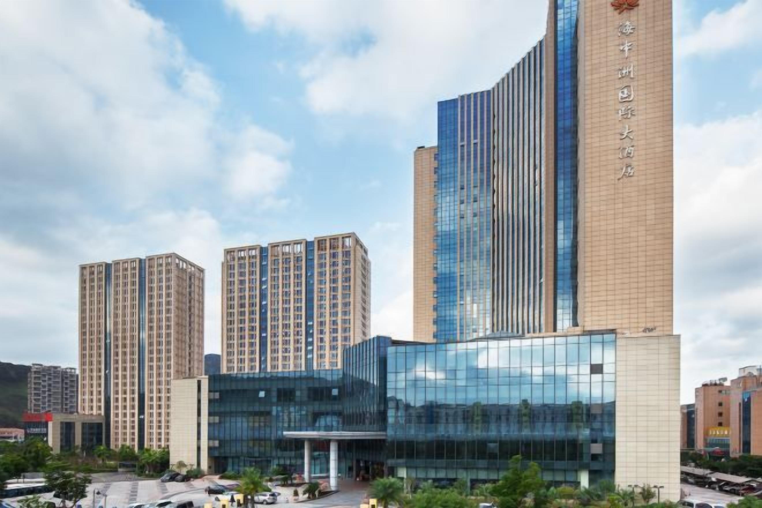 Een modern hotel met een glazen gevel, meerdere hoge gebouwen op de achtergrond en een parkeerplaats voor.