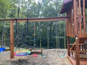 A wooden swing set with a trampoline and a rope swing in a wooded area.
