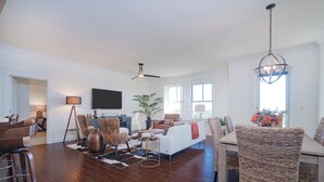 A spacious living room with a large sofa, a dining table, and wicker chairs. There is a ceiling fan, a flat-screen TV, and a potted plant.