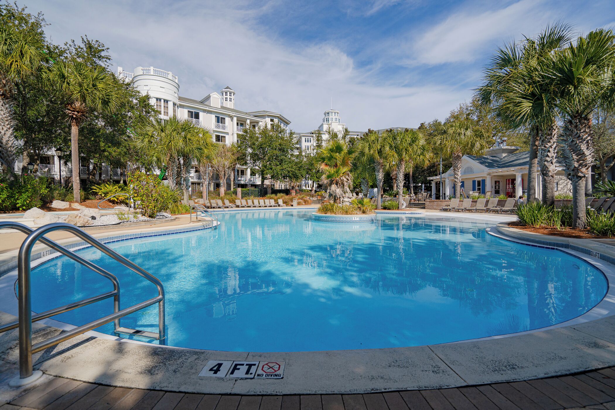 A large, clear swimming pool surrounded by palm trees and a building with a white facade.