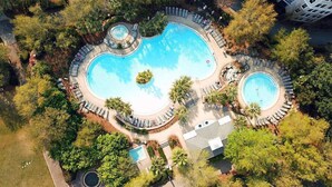 An aerial view of a resort with a large central pool, smaller pools, and lounge areas surrounded by dense greenery.