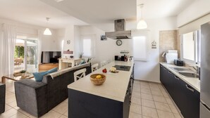 A modern kitchen with a central island, a dining area with a sofa, and a view of the outdoors. - Protaras Villa Athamas Blue (Protaras)