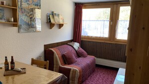 Een knus vertrek met een houten tafel, een zachte bank, een raam met gordijnen en een wandplank vol boeken.