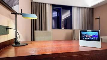 A hotel room with a wooden desk, a digital screen, a lamp, and a view of the outside.