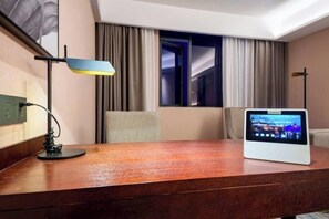 A hotel room with a wooden desk, a digital screen, a lamp, and a view of the outside. - Boyi Hotel (Foshan)