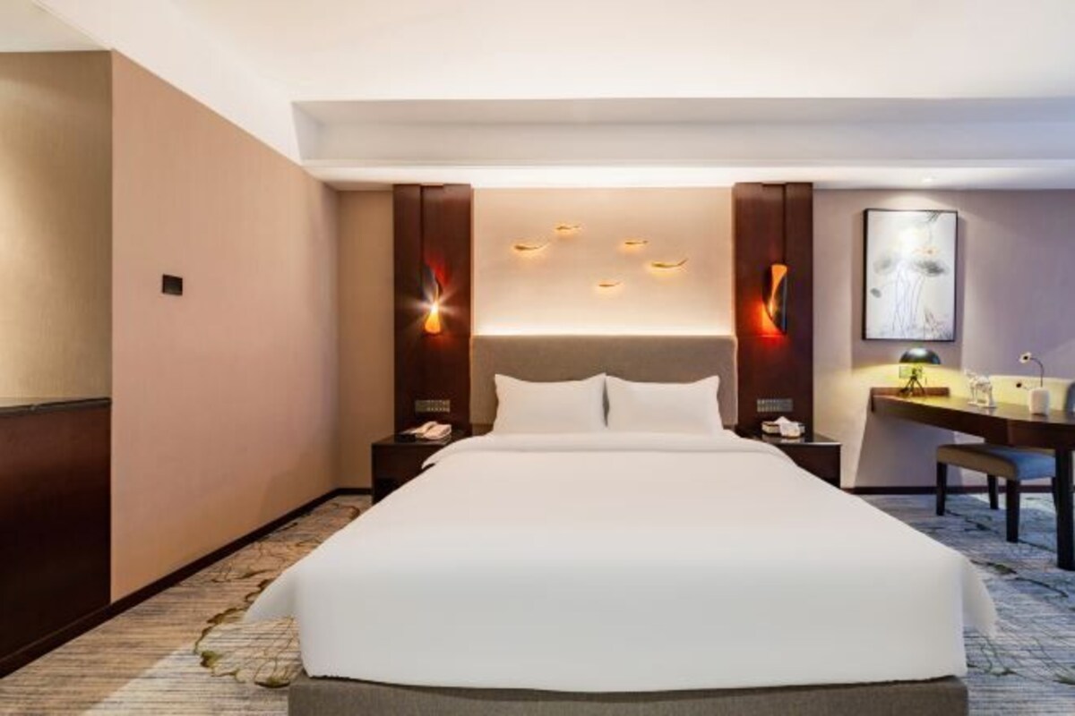 A hotel room with a large bed, a desk, and a chair. There are wall sconces and a painting on the wall.