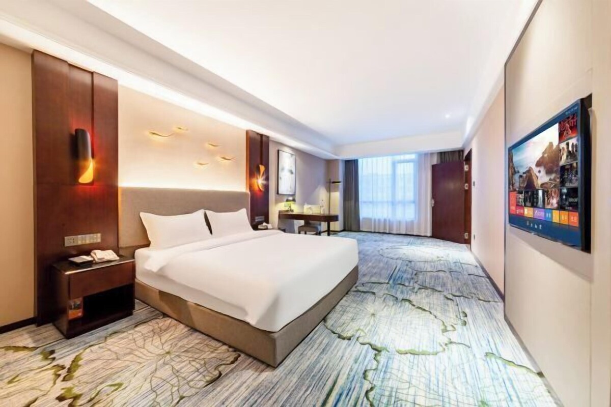 A hotel room with a large bed, a desk, a chair, a nightstand, and a wall-mounted TV.