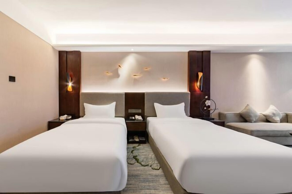 A hotel room with two beds, a sofa, a nightstand with a phone, and a wall-mounted light fixture.