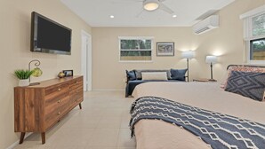 Un dormitorio con cama, cómoda, ventilador de techo, televisor, ventana y un cuadro en la pared.