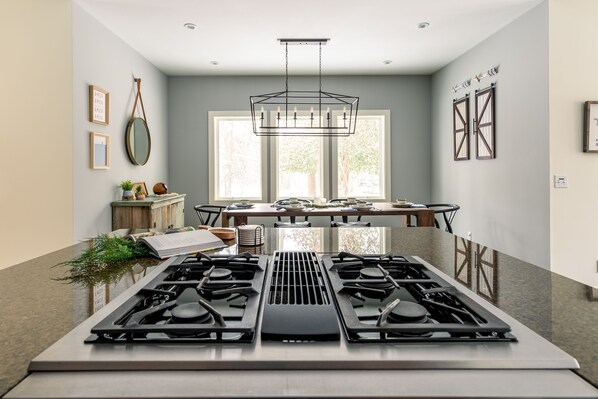 A modern kitchen with a central gas stove, large windows, and a dining area with a table and chairs.