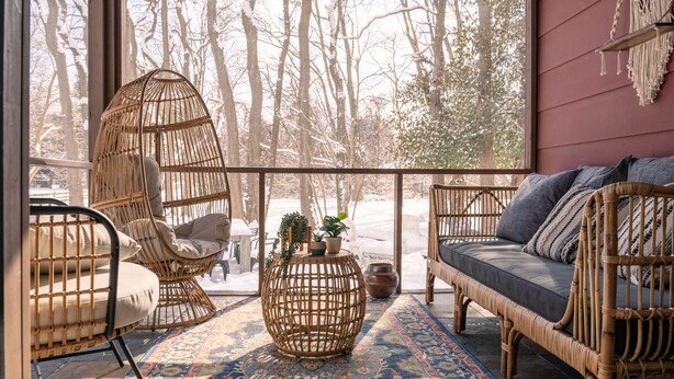 A cozy room with wicker furniture, a patterned rug, and a view of trees through large windows.