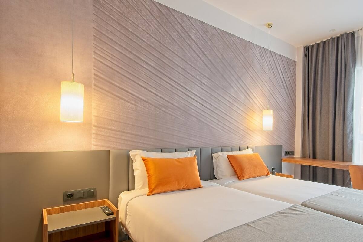 A hotel room with two beds, a wooden nightstand, a desk, and a large window with curtains.