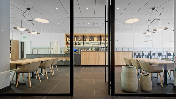 Een moderne eetruimte met lange tafels en stoelen, een centrale bar met flessenrekken, en uitzicht door glazen deuren.
