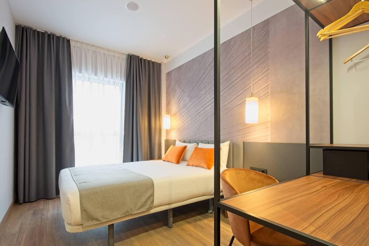 A hotel room with a bed, a desk, a chair, a TV mounted on the wall, and a window with curtains.